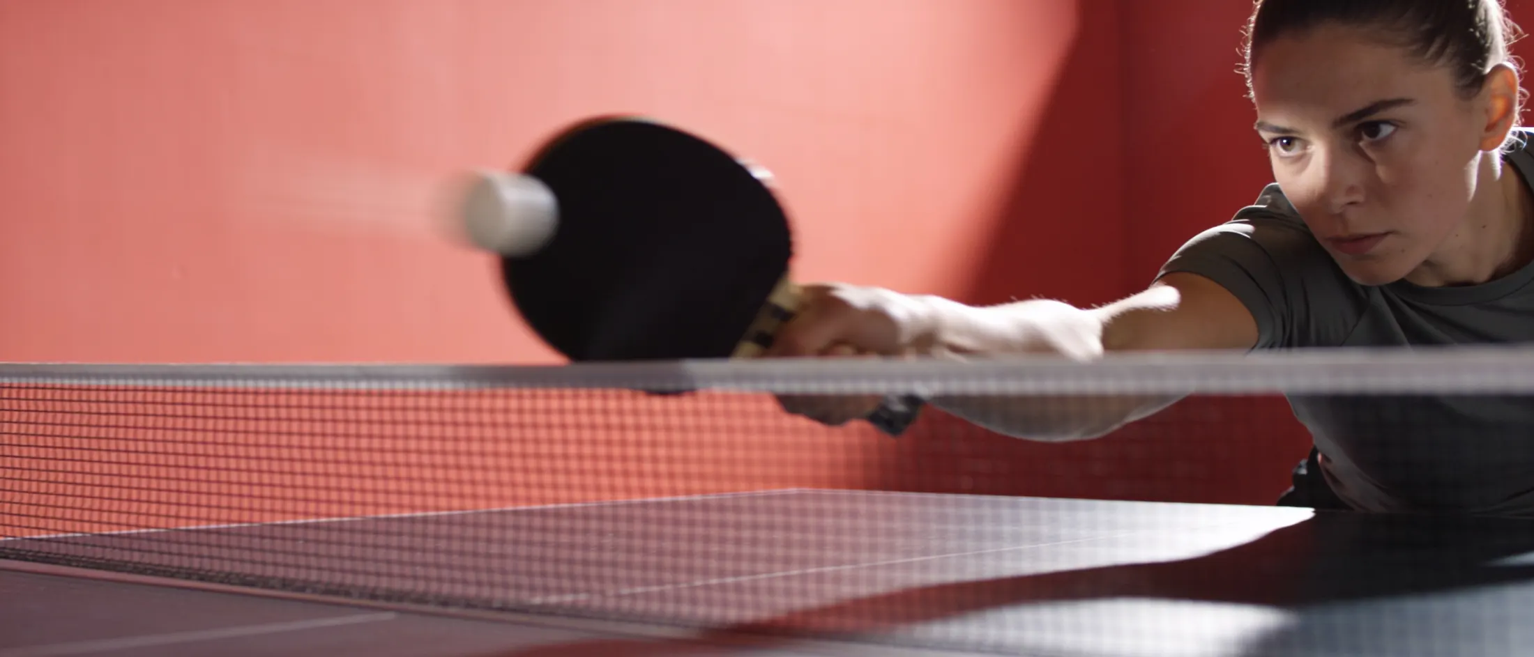 A player driving a forehand at a table tennis table under coral light.
